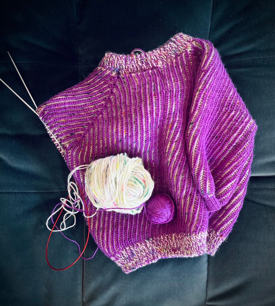 A toddler sweater with one sleeve unfinished on knitting needles. The two color brioche is a bright purple as the main color with a hand painted yarn with bright speckles as the second yarn.