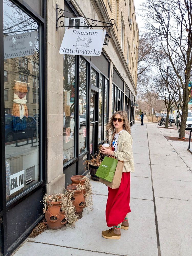 Thirty something in a red skirt, clogs, hand knit cream sweater and rad radishes socks looking very wind blown with shopping bags standing in front of Evanston Stitchworks shop. 