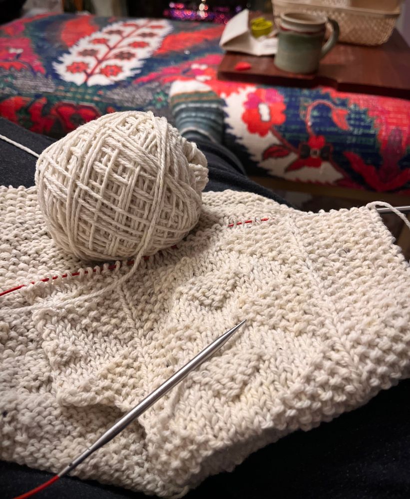 A cream colored ball of yarn on a textured front panel of a hand knit cardigan on knitting needles resting. In the background an ottoman with a mug and fire in a fireplace. Ravelry pattern Esther Jacket #petiteknit