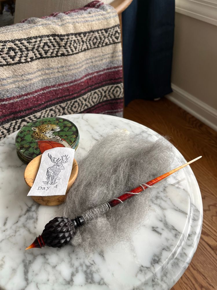 Woodland dragon egg spindle resting on marble table in a small pile of grey wool fiber. Melly Knits True North day 4 moose tag lays beside it.
