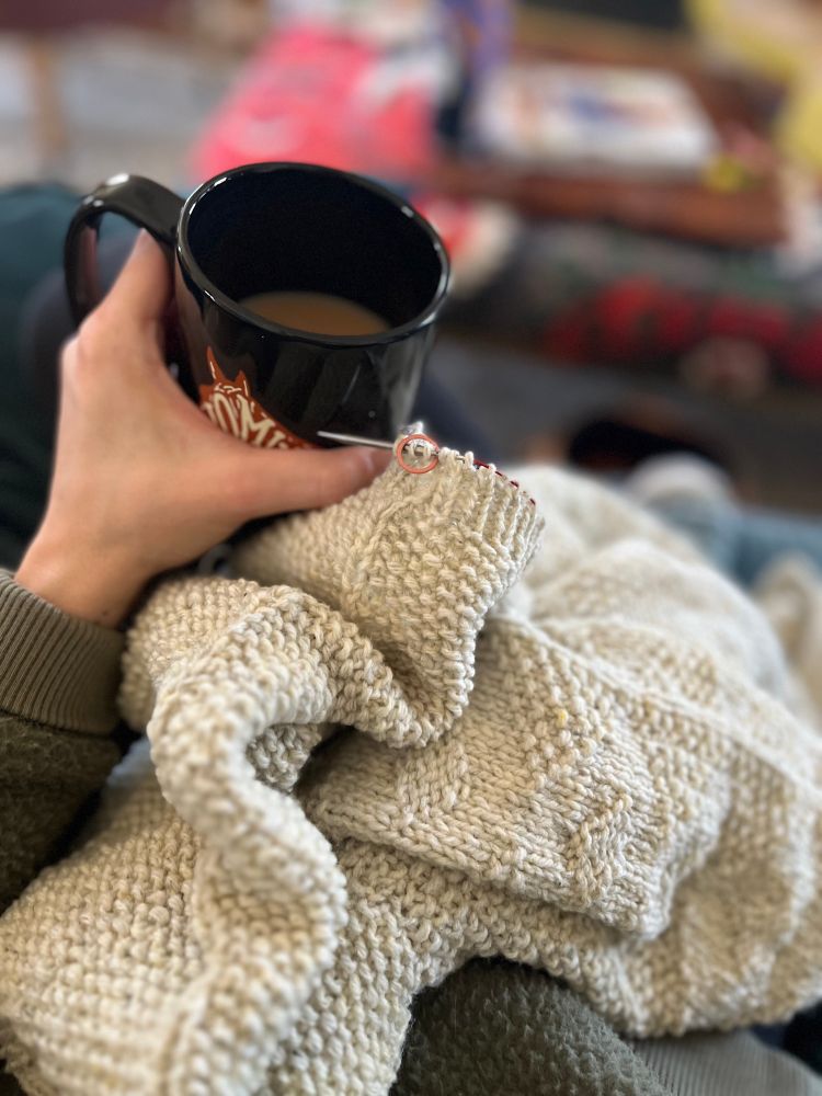 Sweater on knitting needles with a hand holding a cup of tea.