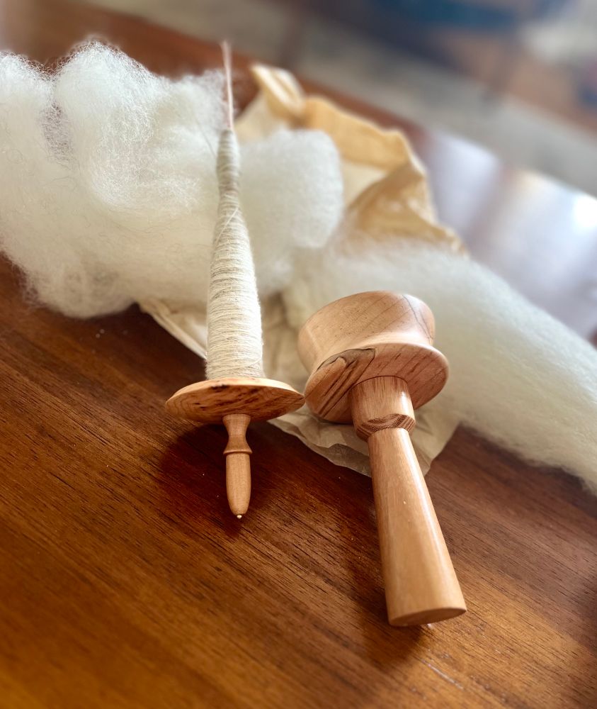 On a wooden coffee table an Enid Ashcroft high worl support spindle in pippy yew is laying next to a lapbowl by Visount Woodworking on a pile of white fluff. Fluff is day 6 Melly Knits advent BFL and alpaca blend.