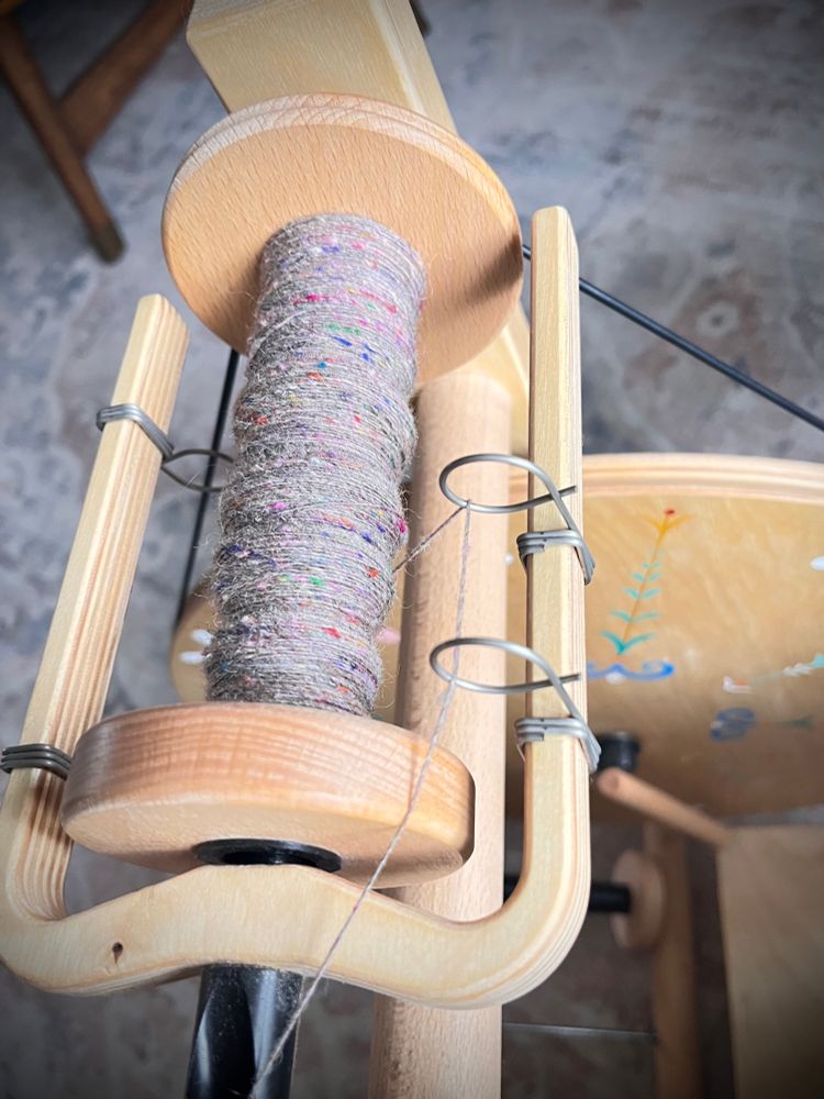 A spinning bobbin 🧵 on a Louet S17 spinning wheel 1/4 loaded with a grey with brightly speckled silk spun wool single.