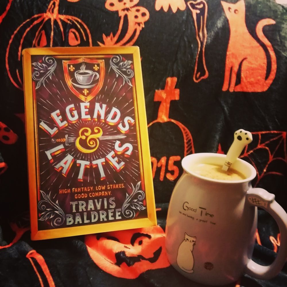 "Legends and Lattes" by Travis Baldree propped up against a backdrop covered with a black blanket with orange spooky pictures on it like cats, pumpkins etc. next to the book is a purple coffee mug with a cat on it