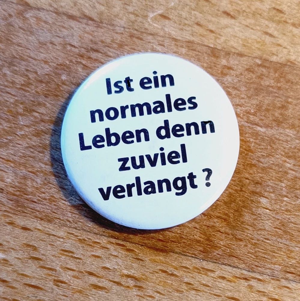 Nahaufnahme: Kleiner weißer Button mit dem Text "Ist ein normales Leben denn zuviel verlangt?" liegt auf einem Holztisch.