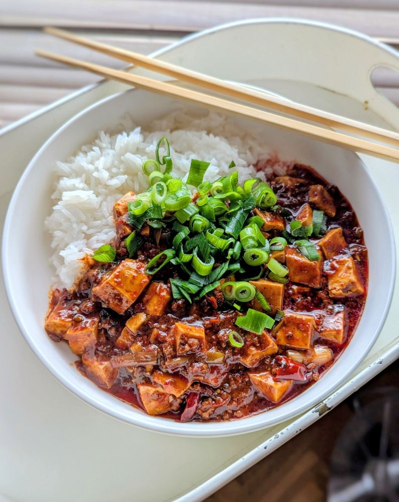 Photo d'une assiette creuse blanche contenant du riz et du lapin tofu bien rouge avec de la cebette fraîche. Deux baguettes sont posées sur le plat. 