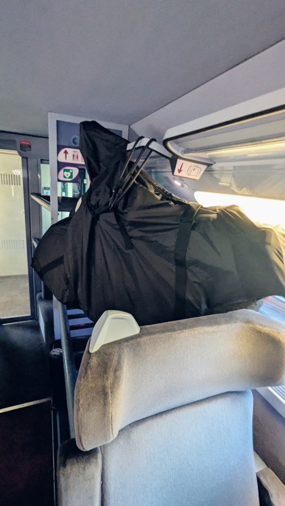 Mon vélo, démonté dans une housse noire. Il est suspendu au-dessus d'un emplacement pour bagages et arrimé avec des tendeurs 