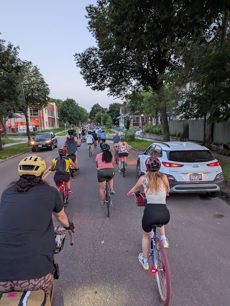 Group of cyclists riding down a residential city street at dusk during a community bike ride. We see helmets, trees, and parked cars.