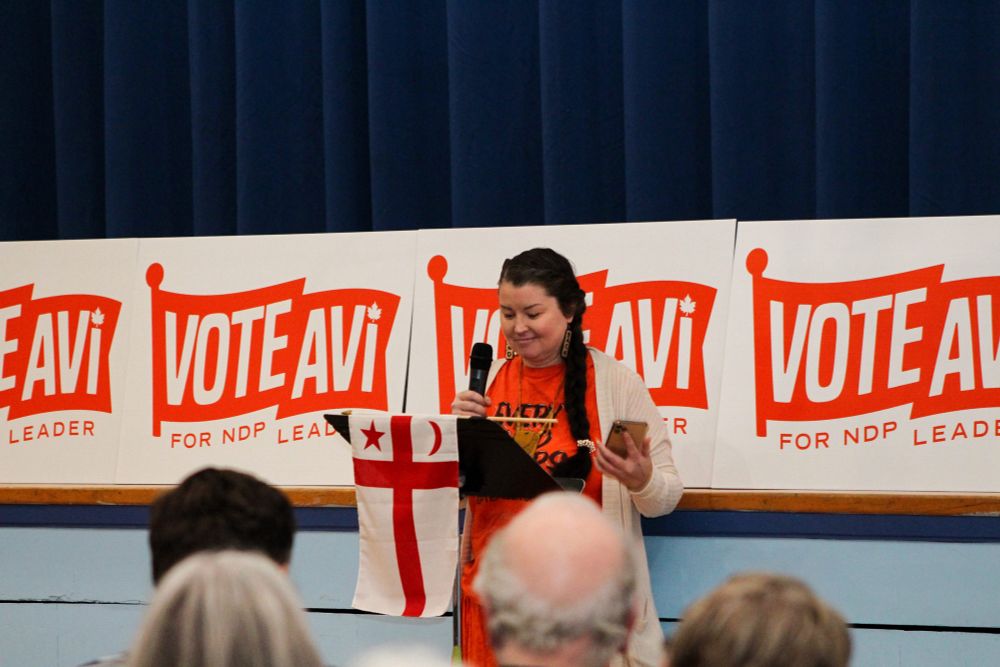 Michelle Paul speaking at Avi Lewis’s, in front of “Vote Avi for NDP leader” signs. 