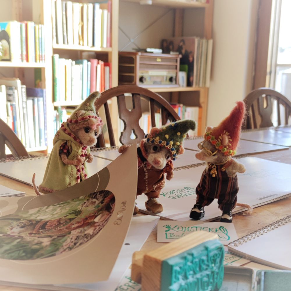 Three felted and clothed mice stand on a table full of calendars. A stamp lies nearby, and they lift the cover of one calendar, peering curiously underneath.