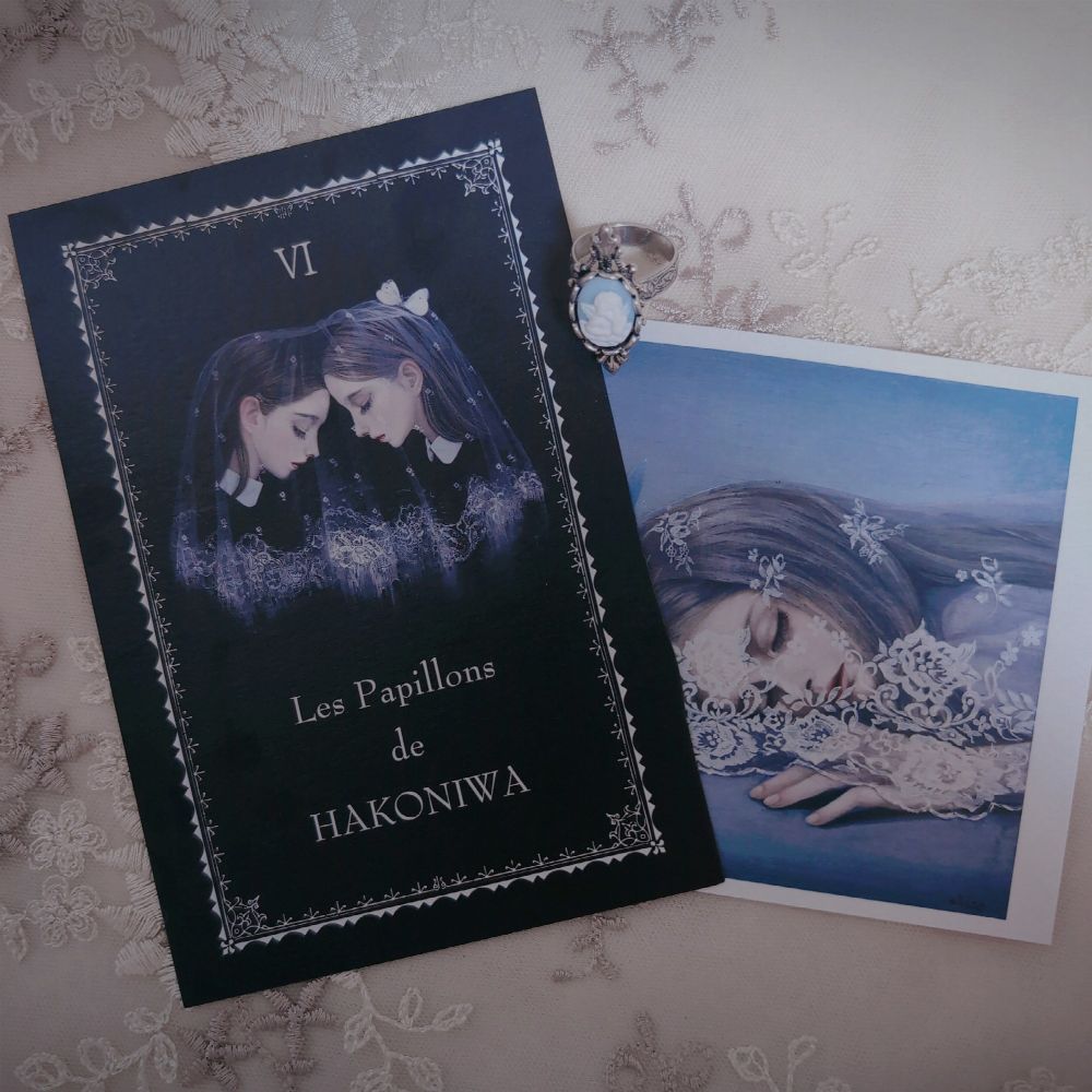 黒い表紙に向かい合う少女のイラストと「Les Papillons de HAKONIWA」の文字、隣にレースをまとい眠る少女の青い絵、カメオ風の指輪が添えられている。