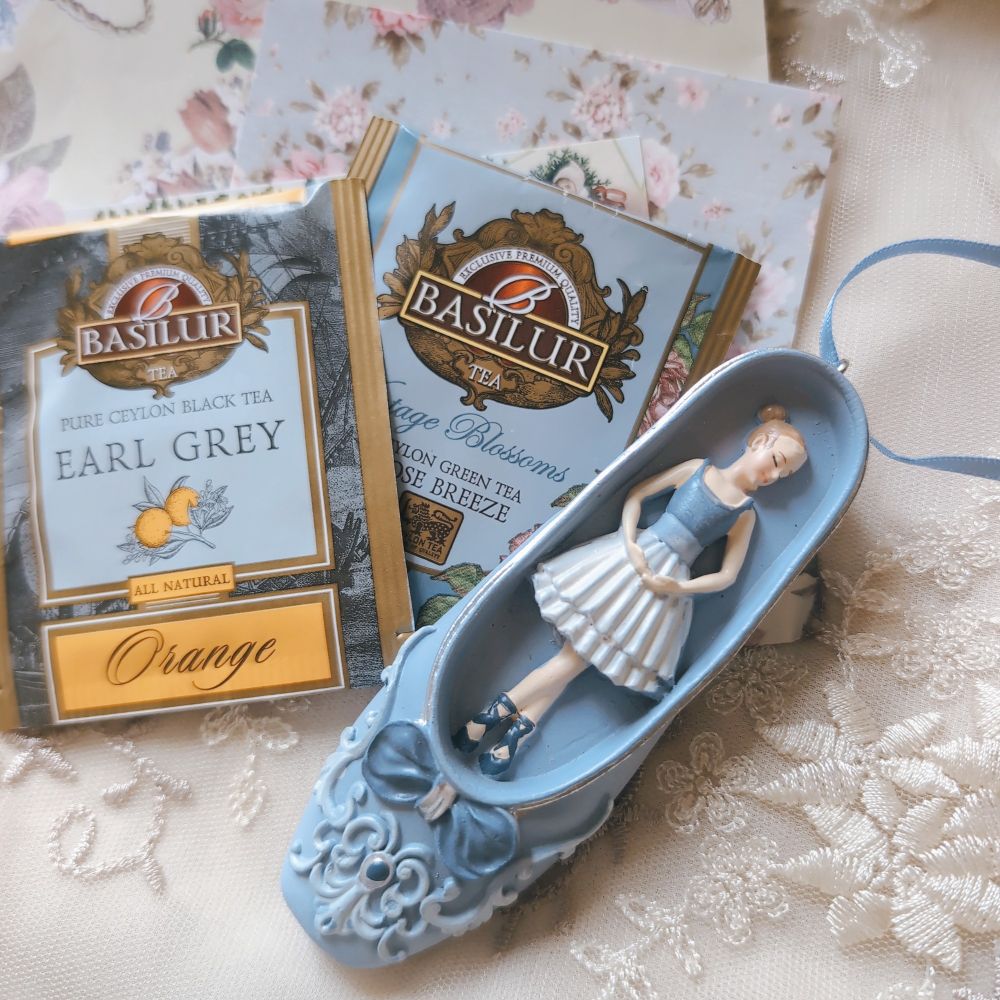 レースの布の上に置かれたオーナメント。
ブルーのリボンやレース風の装飾、宝石風の装飾がついた青いトウシューズの中に、バレリーナの人形が足をクロスさせ手はアン・バーのポーズを取っている。
バレリーナはシニヨンに髪を結い、ブルーと白のロマンティックチュチュ、ブルーのトゥシューズ姿。
後ろにはバシラーティーのアールグレイとオレンジブロッサムのティーバッグ、アンティーク調の薔薇模様の紙が添えられている。