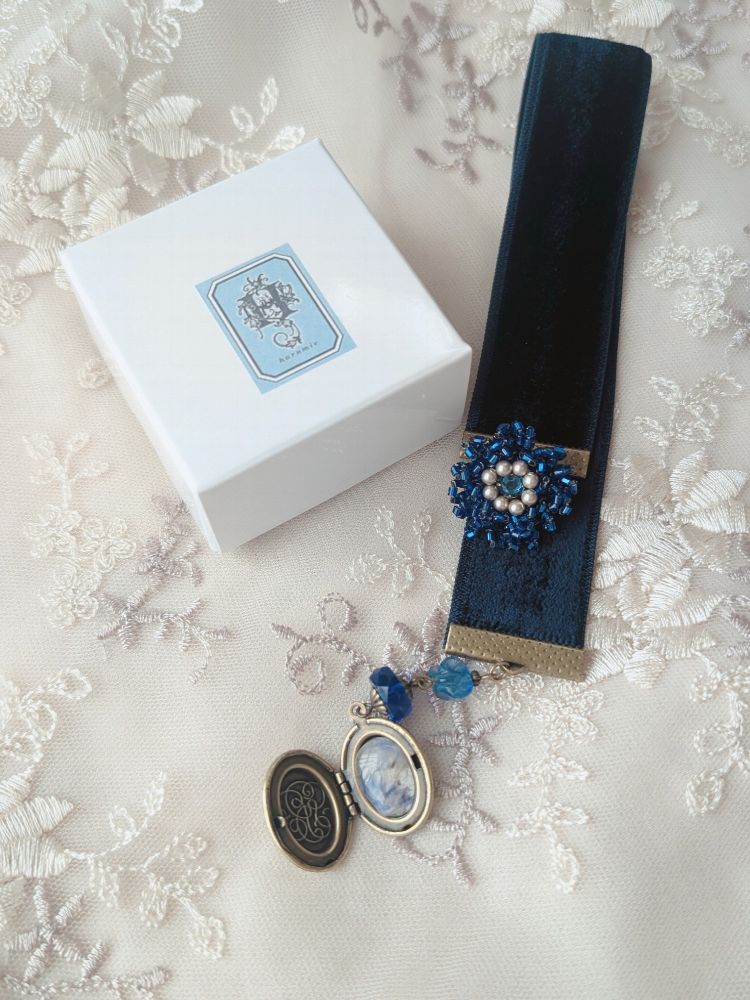 白いレース地の上に、青いビーズの花飾りが付いた黒いベルベットのブックマーカーと、青い石とロケットチャームが付いたアクセサリー、小箱には「hermine」と書かれたラベルがある。