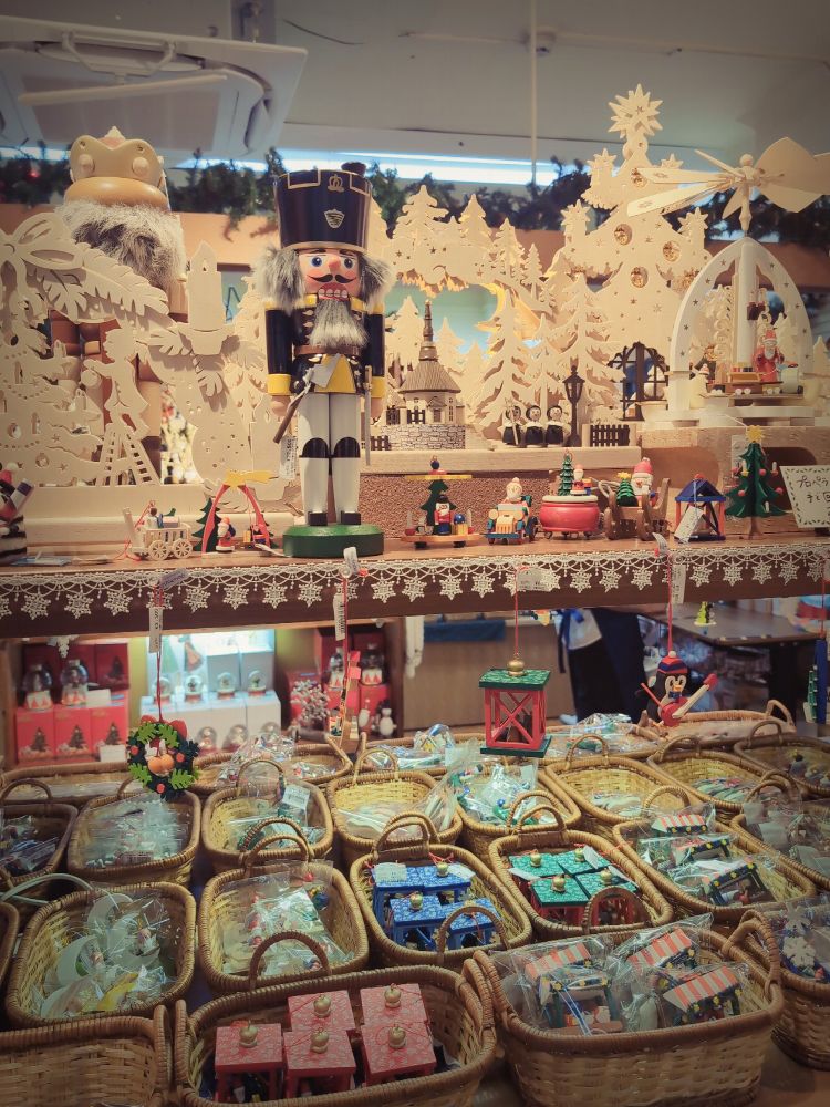 クリスマス装飾が施された店内で、くるみ割り人形や木製オーナメント、ミニチュアの家などがカゴに並ぶドイツ風の雑貨売り場の様子。