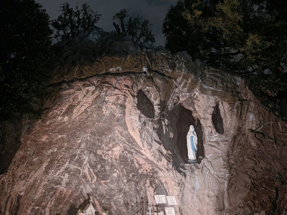 岩肌に彫られた洞窟の中に白と青の衣をまとった聖母マリア像が立ち、周囲には木々と「アヴェ・マリア」などの文字が書かれた案内板がある夜の風景。