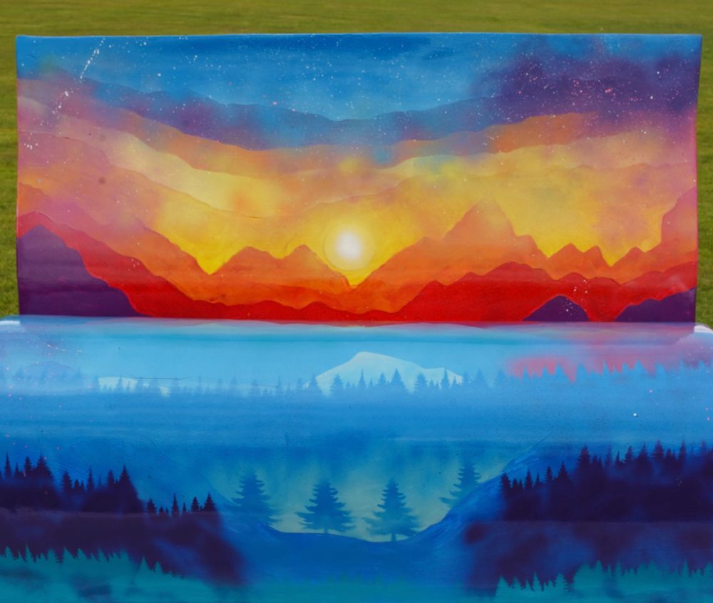 Brightly coloured reading bench. The top is a fiery sun in mountains and the bottom is misty woods and pine trees.