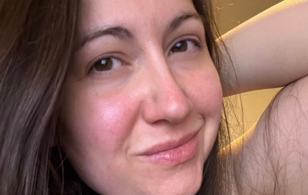 A closeup selfie of a woman with brown hair and no makeup, smiling for the camera 