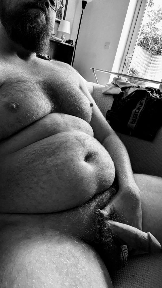 A black and white photo of a hairy bearded man sitting nude on a couch. He is grasping his erect penis