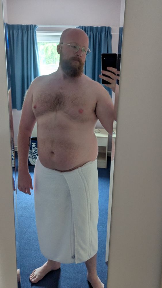 A bald ginger bearded man takes a selfie in a mirror while wearing a towel around his waist