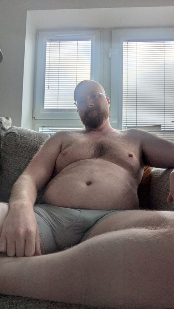 A man reclines on a couch in front of a window with grey skies. He is wearing grey boxer briefs and his bulge is clearly visible 