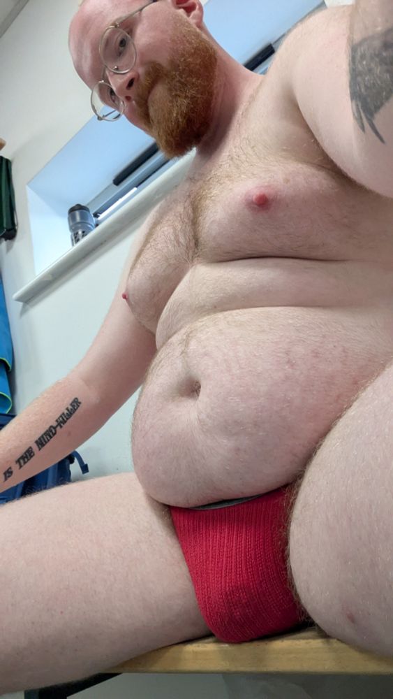 A red bearded man wearing glasses and a red jockstrap takes a low angle selfie sitting in a changing room. 
