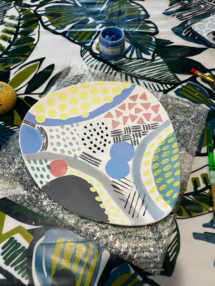 picture of a misshaped white plate with round edges laying on top of bubble wrap on a table with a leaf pattern. The plate is painted with a variety of geometric shapes in different colors 