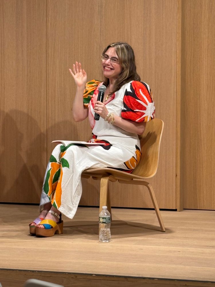 Maris Kreizman, author of I Want to Burn this Place Down, waving to crowd at her book launch event at the Brooklyn Public Library. She wears a gorgeous long dress with bright flowers against a white field, and multi- colored shoes. 