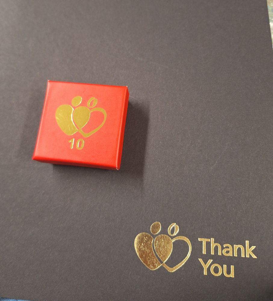 A small square red box with the blood donation service logo on it and the number 10 is sitting on a dark coloured certificate folder with the same logo and the words "thank you".