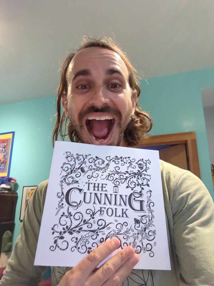 Colin is holding up a copy of The Cunning Folk, a beautifully illustrated new TTRPG book, wearing an extremely excited face.