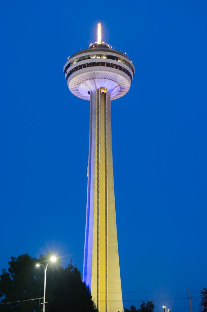 A tall concrete tower with the top wider than the base.  The sky is just after sunset.  There are trees and a street light at the lower left of the image.  