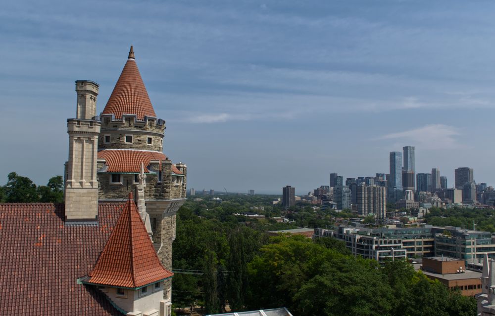 A round castle like structure to the left of the image with a red cone shaped roof.  To the left  is a city skyline.  The sky has some clouds but is mostly blue.