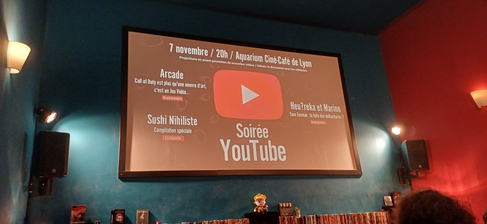 Photo de l'Aquarium Ciné-Café. Un écran diffuse une image avec les textes suivants :
Soirée Youtube 

Arcade
Call of Duty est plus qu'une œuvre d'art, c'est un jeu vidéo 

Sushi Nihiliste
Compilation spéciale 

Heu?reka et Marino
Taxe Zucman : la fuite des milliardaires ?