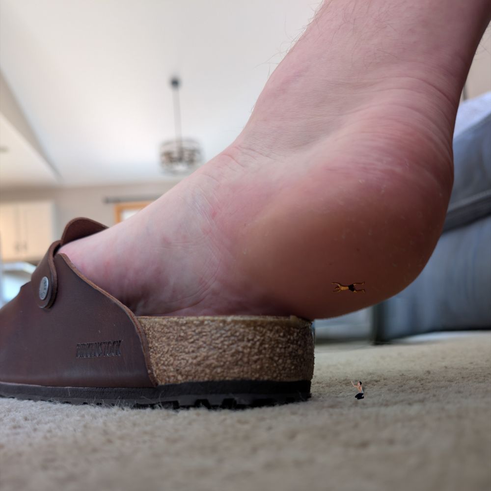 Floor shot of my foot popped out slightly out of a Birkenstock clog, on my heel is a tiny man stuck on, my heel is looming over another tiny man.