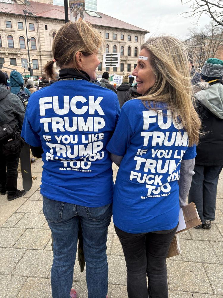 Two women with their backs to the camera and blue t-shirts that say “Fuck Trump If You Like Trump Fuck You Too” on the back