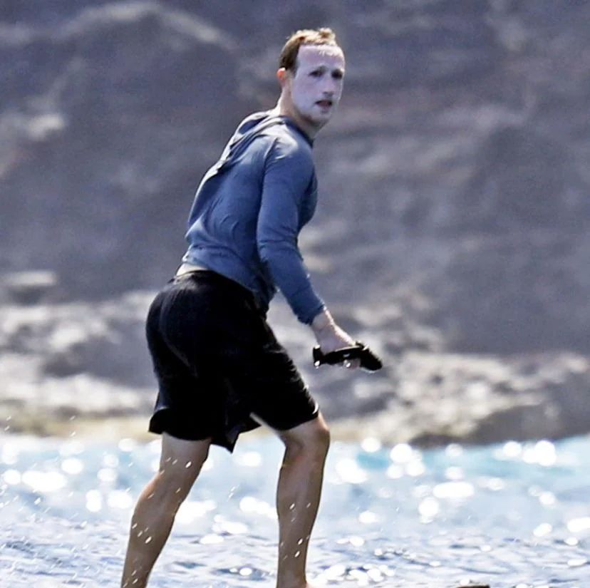 Zuckerberg surfando, de bermuda preta, blusa de manga azul e o rosto tomado por uma grossa camada de creme branco