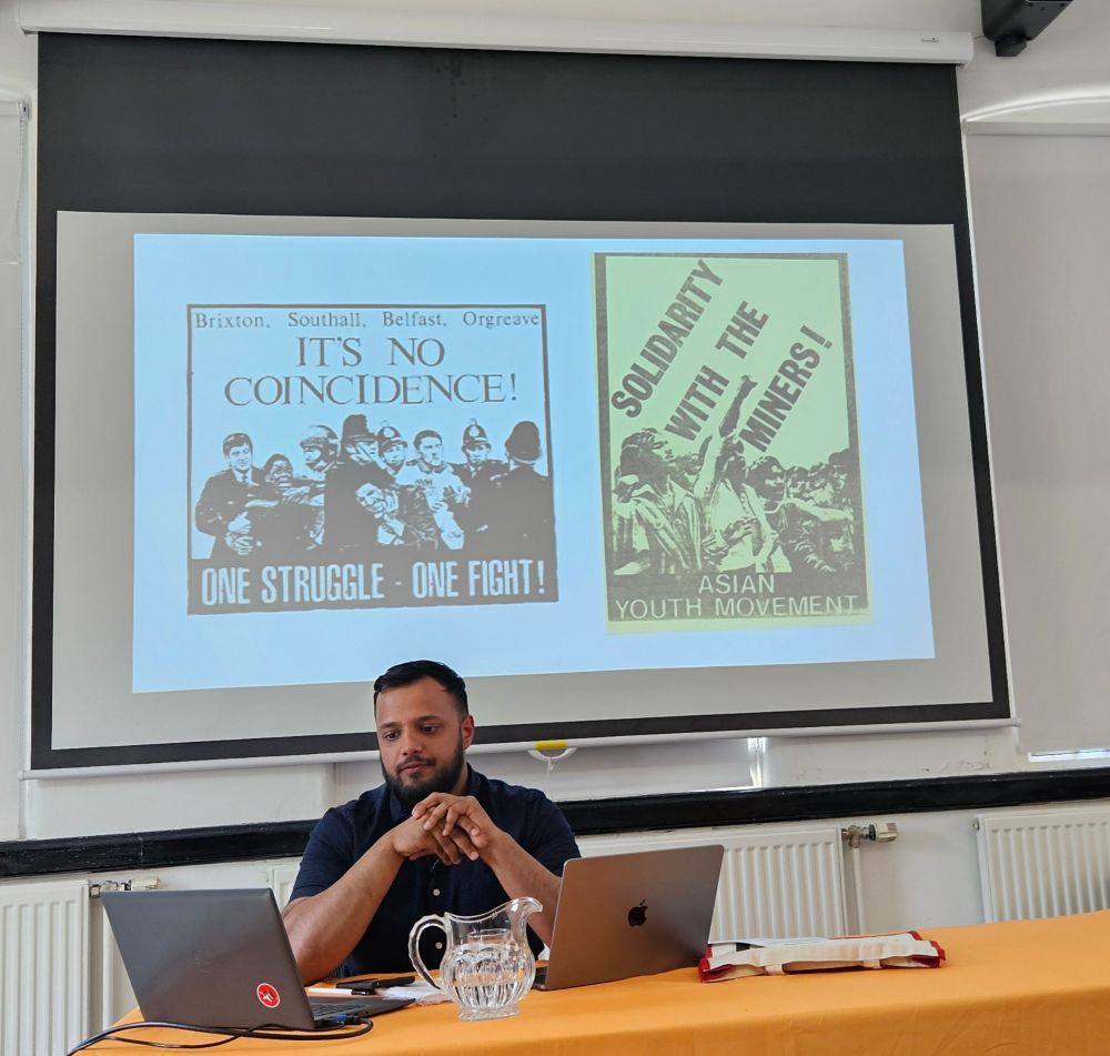 Taj Ali in front of slide two posters one on joint struggles with Orgreave Toxteth Bradford Southall. The other with Asian protesters labelled Solidarity with the Miners