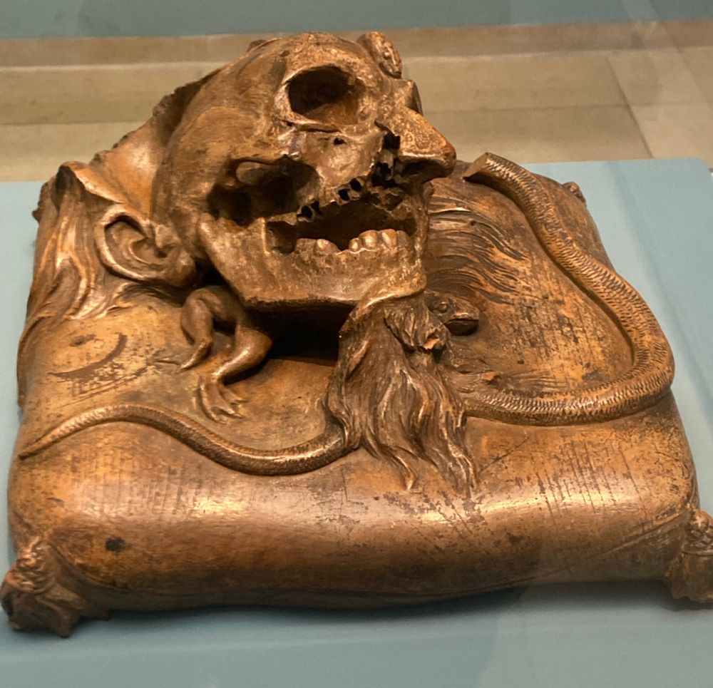 Pearwood carving of a largely decomposed human head (right ear, beard and some hair intact) resting on a tasselled cushion. A snake (its head broken off) is coiled around the edge