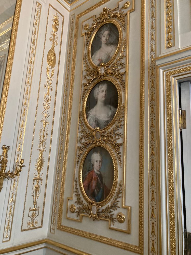 Three oval portraits set into the white woodwork in the corner of a room. Relatively restrained gilt boiseries surrounding them - more neo-classical Louis XVI than rococo