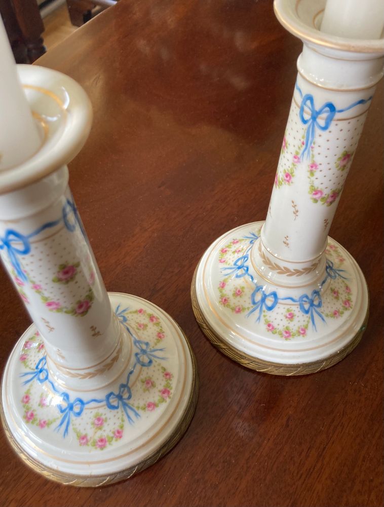 Pair of white porcelain candlesticks with wide bases, rimmed with ormolu. Painted with garlands of pink roses tied with blue ribbons surrounding larger roses amidst gold dots.