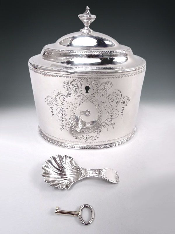 Oval bowl with straight sides. Cover hinged and domed with vasiform finial. On front, bright-cut oval wreath (vacant) surrounded by leafing scrollwork, pendant flowers, and garland. Beading. Spoon: Scallop shell bowl; tapering and bright-cut handle. With key. Both spoon and caddy fully marked including duty stamp. Caddy has duty drawback stamp (in effect, an export stamp).