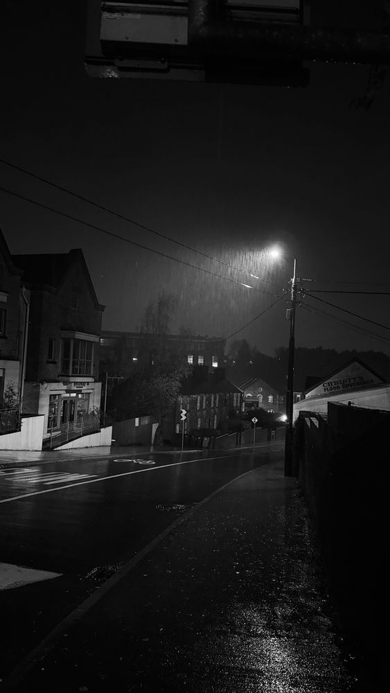 Black and white night photograph of Donnybrook Mills and surrounding cottages in the rain. A streetlight illuminates falling rain and wet pavement, reflecting light along a quiet, empty street lined with old brick buildings and shopfronts. Power lines cut across the dark sky, adding to the moody, atmospheric scene.