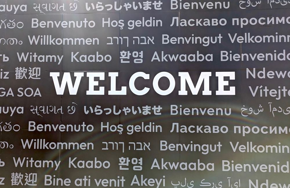 ALT tag:
Welcome sign featuring the word ‘Welcome’ in large white letters surrounded by translations in multiple languages on a dark background.