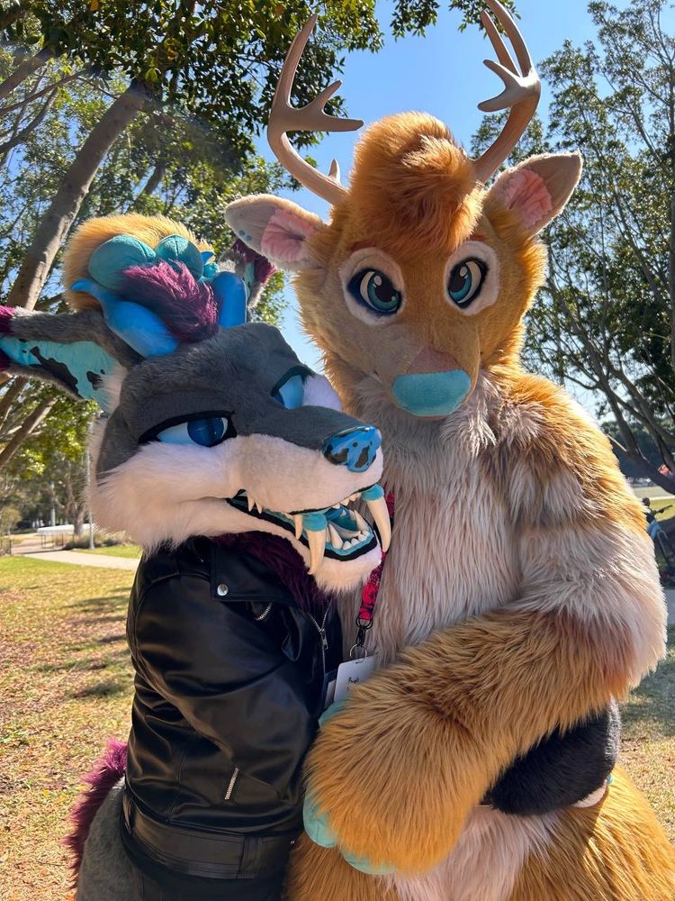 Luka chupacabra on the left, in a partial fursuit with grey, white and purple fur. Blue accents on the horns, ears, eyes, nose and inside of the mouth. He has sharp teeth and is hugging around Tyba’s waist. Tyba is a deer fursuit with light orange-brown fur, with cream markings and blue nose and hoof hands. Hugging Luka in return and petting the top of his head.
