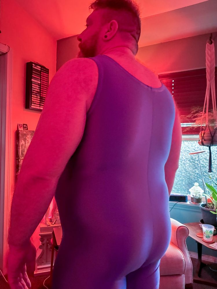 The same man is seen from behind, showing the back of the purple singlet under soft red lighting in a cozy room.