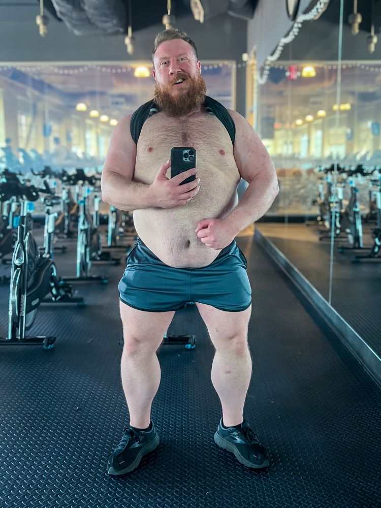 Bearded man with a muscular, thick build posing shirtless in front of a gym mirror. He is wearing shorts and sneakers, with a sweaty, proud expression after a workout.