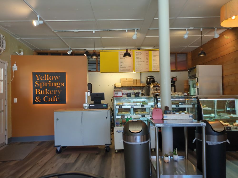 Alt text: Interior of Yellow Springs Bakery & Café, featuring a warm, inviting design with wood accents and modern lighting. The counter area displays pastries and baked goods in glass cases, with a menu overhead. A sign on an orange wall reads "Yellow Springs Bakery & Café" in illuminated cutout letters. Two trash bins and a small cart with succulents and utensils sit in the foreground.
