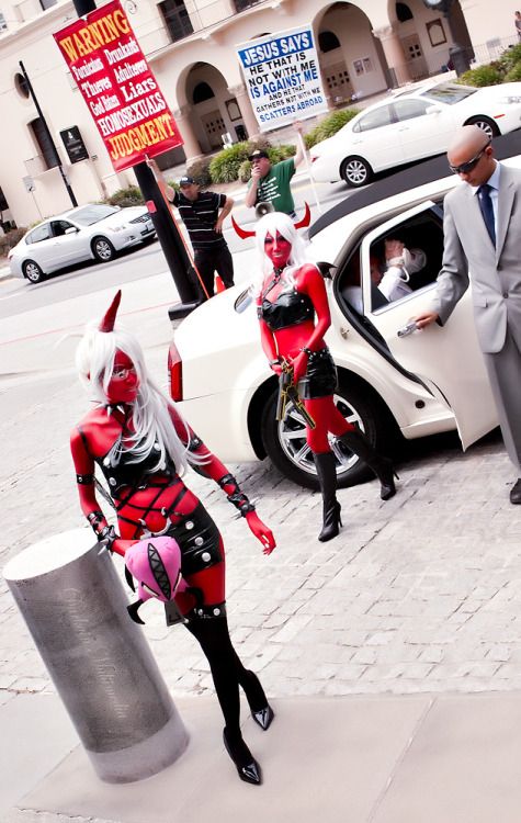 Cosplayers portraying the demon woman characters Scanty and Kneesocks stepping out of a limousine while evangelical protesters hold signs in the background