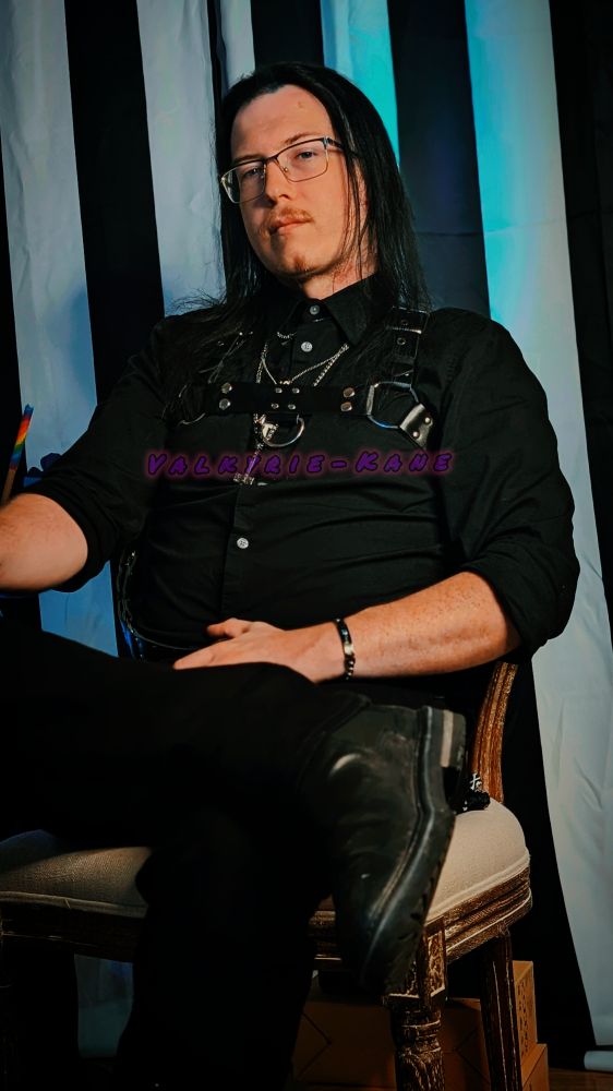 Valkyrie-Kane, with straight hair instead of wavy, sits elegantly in an antique chair. They are dressed in their kink attire, a black harness and black leather boots, with their black shirt and pants.