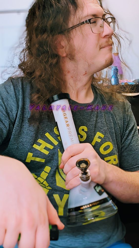 Valkyrie-Kane sits and is holding their bong as they stare off to the right with a pleasant expression on their face.