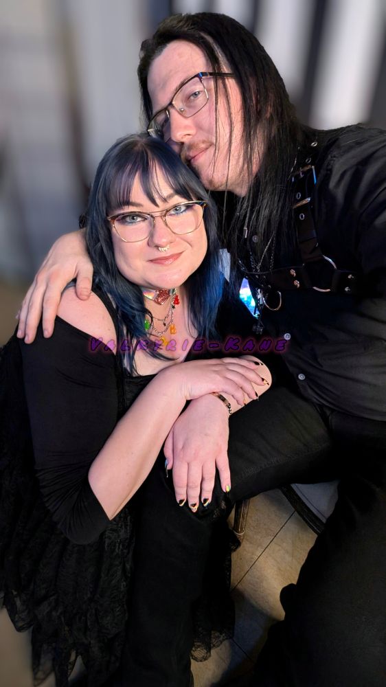 Valkyrie-Kane leans down and arm wraps Celleste as she kneels and rests her arms on their lap.